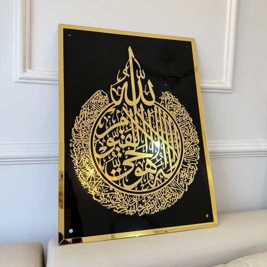Decorative piece with gold Arabic calligraphy on a black background, framed in gold, against a white wall.