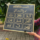 Person holding a black board with gold Arabic calligraphy against a green leafy background