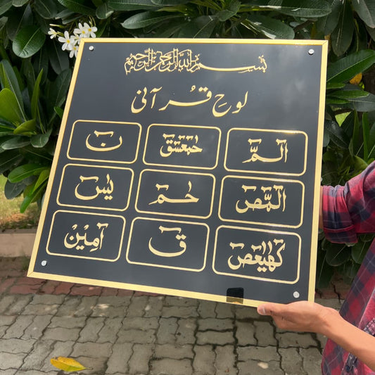 Person holding a black board with gold Arabic calligraphy against a green leafy background